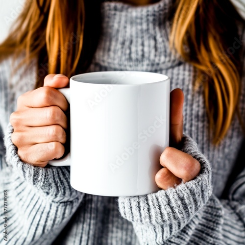 A woman in a sweater holds a mockup of a white mug in her hands. (ID: 777790418)