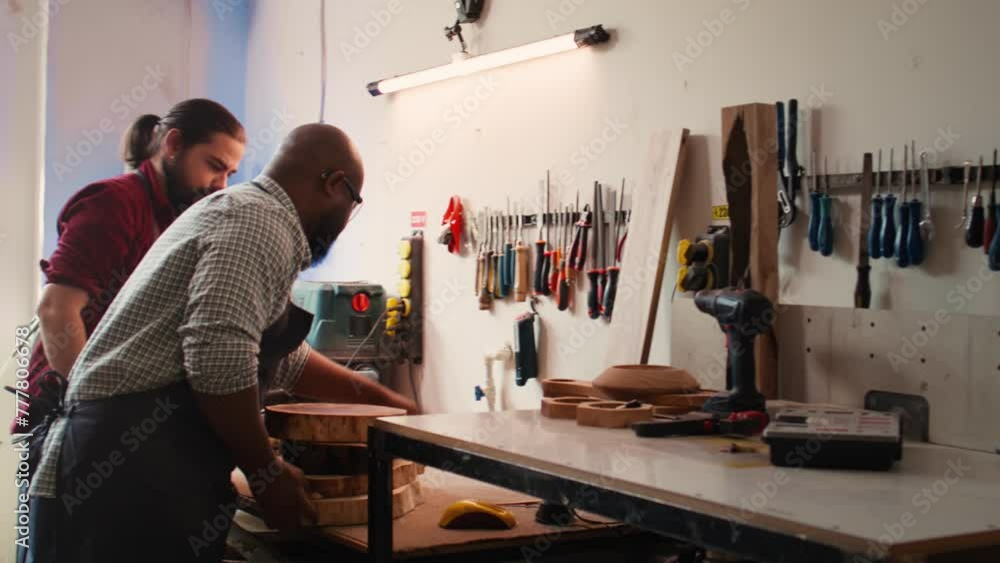 Carpenter selecting tool from rack to use on wood materials, helped by african american coworker. Craftsperson doing quality assurance on lumber pieces pile with BIPOC apprentice, camera B Carpenter selecting tool from rack to use on wood materials, helped by african american coworker. Craftsperson doing quality assurance on lumber pieces pile with BIPOC apprentice, camera B