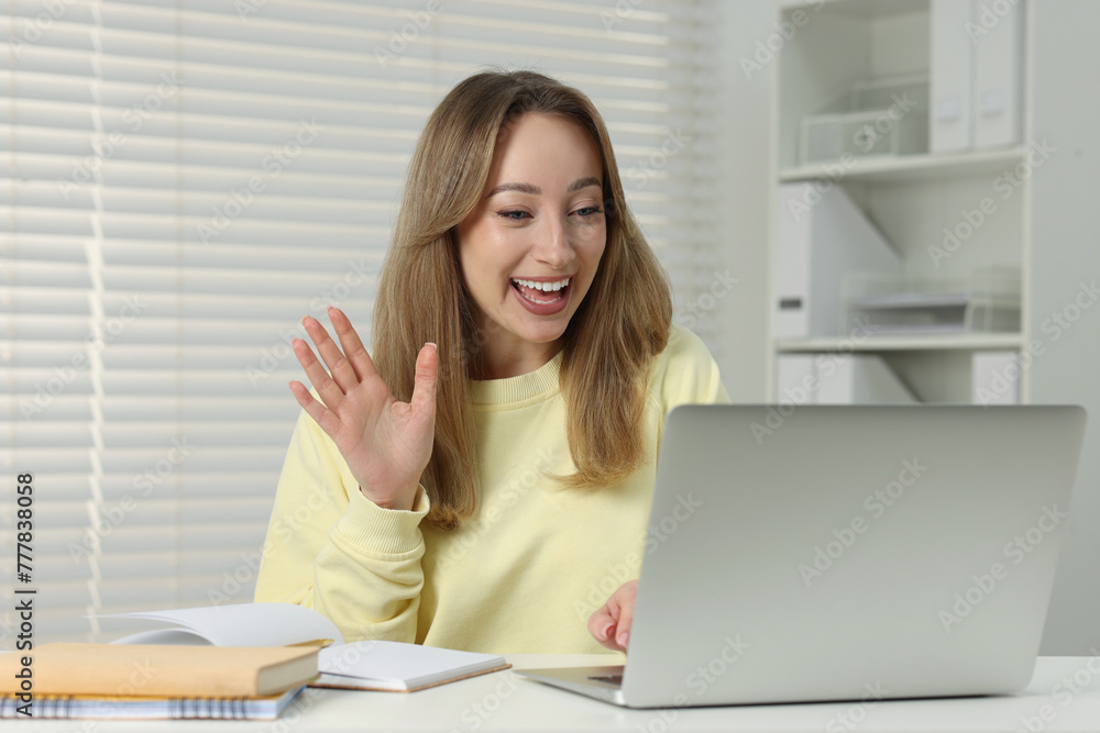 Young woman waving hello during video chat via laptop at white table indoors Young woman waving hello during video chat via laptop at white table indoors