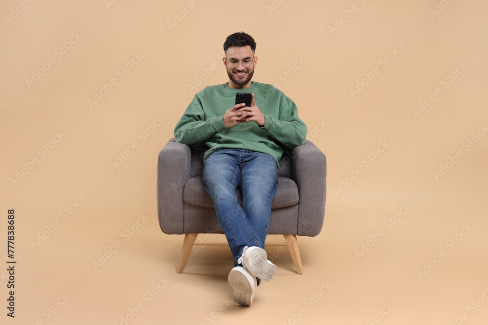Happy young man using smartphone in armchair on beige background Happy young man using smartphone in armchair on beige background