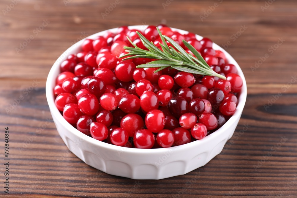 Fresh ripe cranberries and rosemary in bowl on wooden table, closeup Fresh ripe cranberries and rosemary in bowl on wooden table, closeup