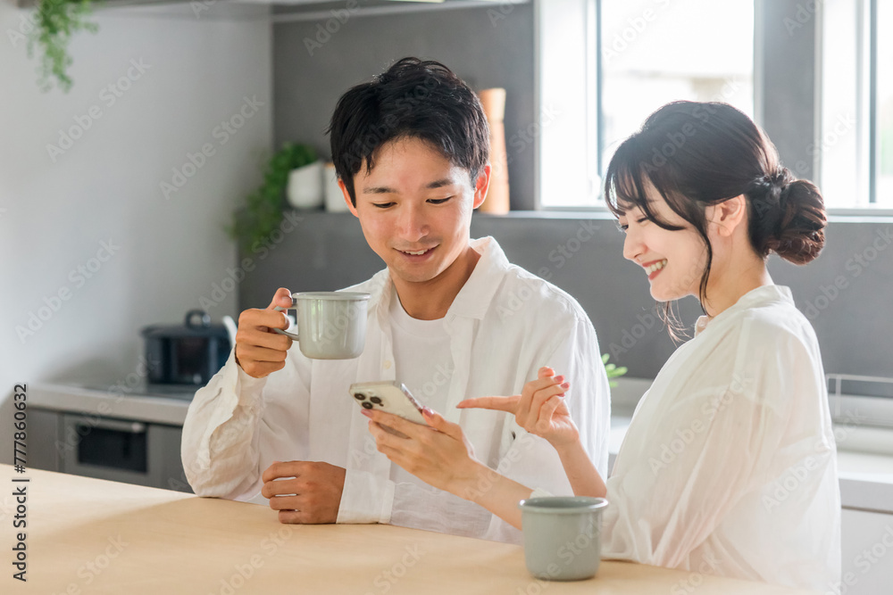 リビング・ダイニングでスマホを見ながらティータイムを過ごす仲良しの若い夫婦・カップル
