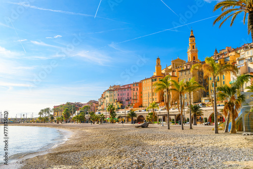 The sandy Plage des Sablettes beach and promenade, with the colorful old town and the towers of Basilica of Saint Michel behind, along the French Riviera at Menton, France.