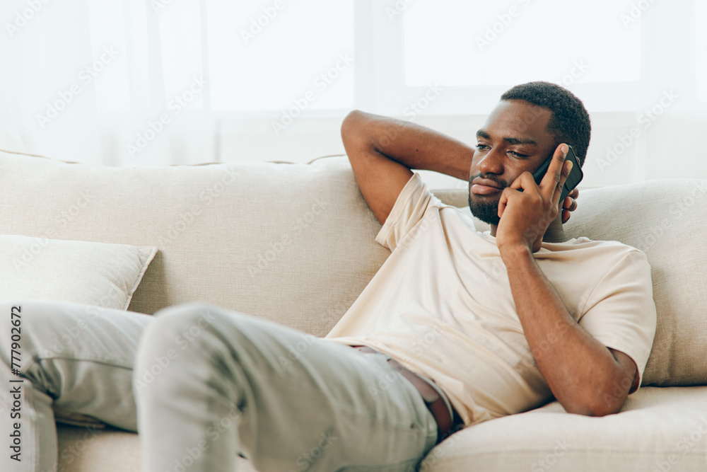 Happy African American man sitting on a black sofa, using his mobile phone for a video call The modern apartment creates a comfortable backdrop as he confidently chats and relaxes With a smile on his