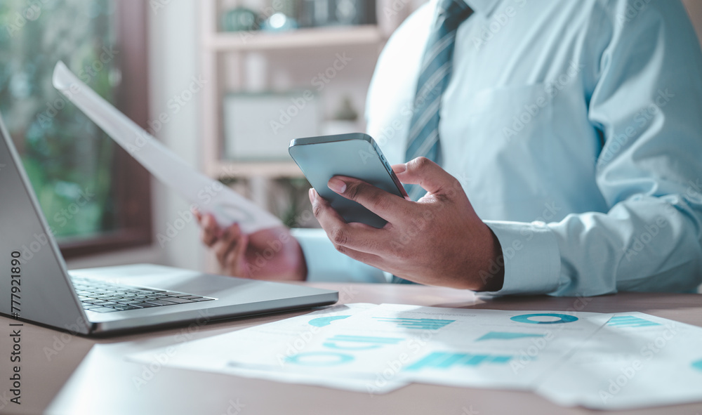 chart, document, graph, management, paperwork, plan, report, research, debt, income. A man is sitting at desk with a laptop and smartphone. and he is focused on something. chart, document, graph, management, paperwork, plan, report, research, debt, income. A man is sitting at desk with a laptop and smartphone. and he is focused on something.