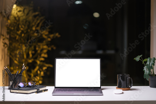 Laptop with blank screen coffee cup and potted plant on white table in modern dark office at night.