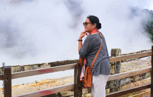 Rear view: A woman standing near a wooden fence looks at one side with a smile, the background is white sm coming out of the Sikidang Crater photo