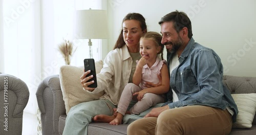 Loving parents with little 4s daughter using smart phone on weekend at home, sit on couch look at gadget screen, making videocall enjoy remote talk to family, having fun enjoy new mobile application photo