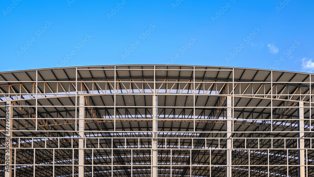 Large metal industrial factory building structure with corrugated steel curve roof and skylights in construction site against blue sky background, front view with copy space 