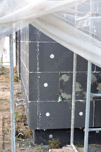 A wall of a house covered with EPS graphite polystyrene boards for thermal insulation, a scaffolding and building safety scaffolding net, anchors, an exterior wall mounted water faucet © E-lona
