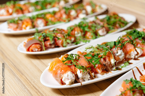 Smoked Salmon Rolls with Cream Cheese and Arugula. Plates of smoked salmon rolls filled with cream cheese, garnished with arugula and a balsamic glaze, arranged on a wooden table.