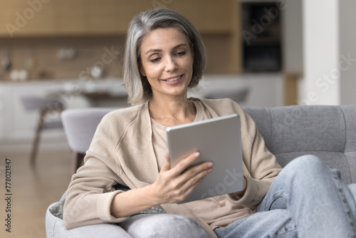 Smiling attractive middle-aged woman relax sit on cozy couch, hold digital tablet, using new application for hobby, browse internet, read news, buy through commercial websites, enjoy free time at home