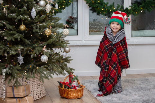 Happy little girl in a santa claus hat wrapped in a warm plaid blanket waiting for santa claus to come.