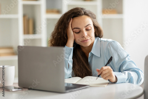 Sad woman looking annoyed and stressed, sitting at table in front of laptop, thinking, feeling tired and bored, having lack of inspiration