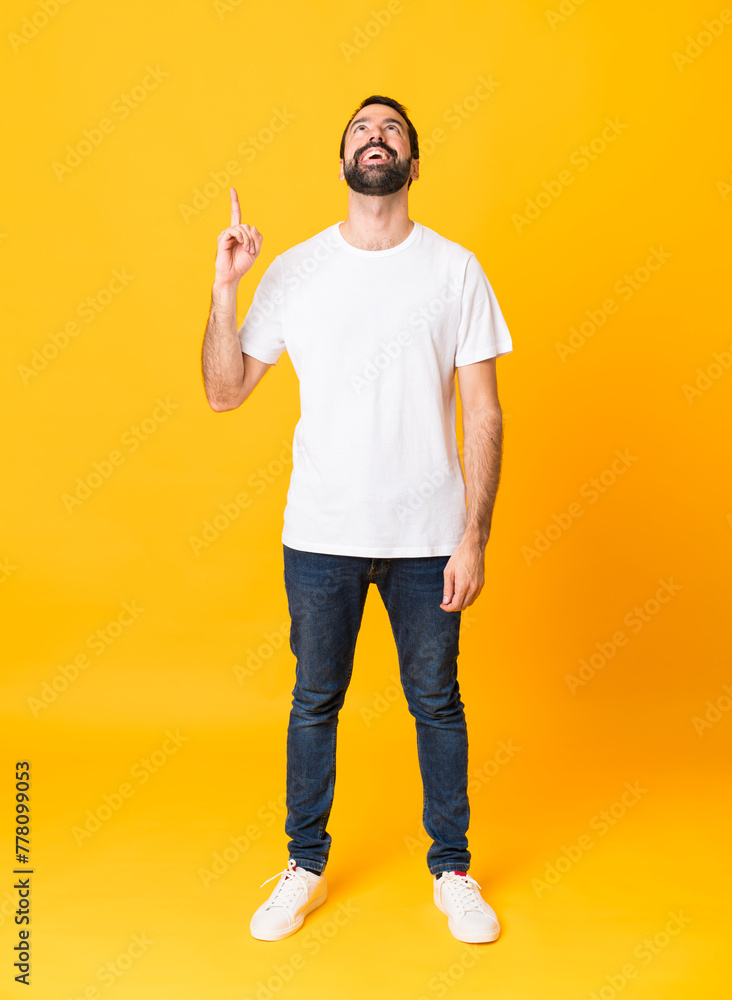 Full-length shot of man with beard over isolated yellow background pointing up and surprised Full-length shot of man with beard over isolated yellow background pointing up and surprised