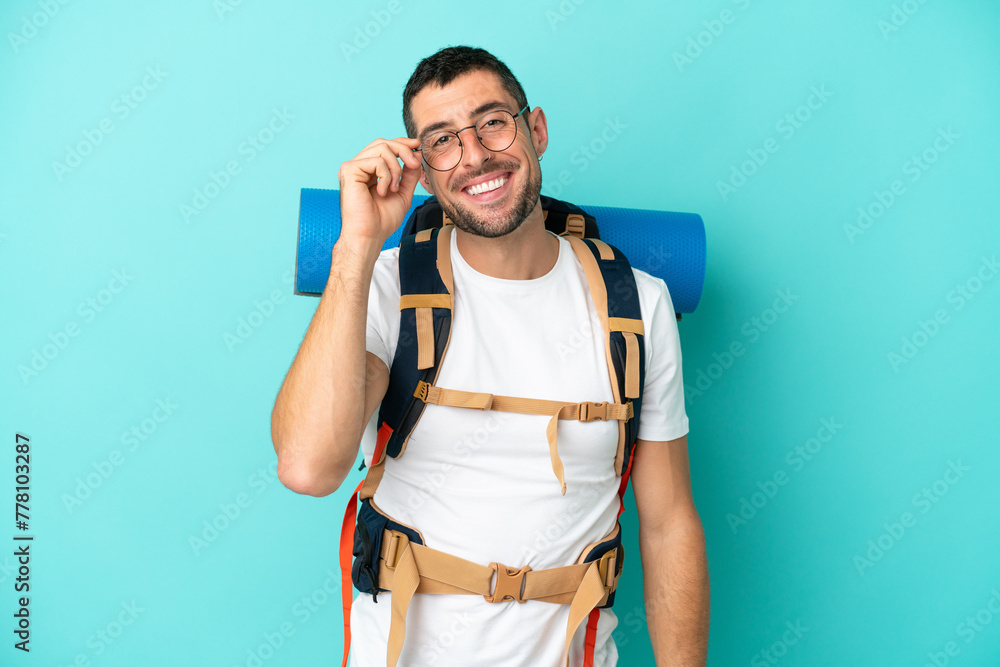 Young mountaineer caucasian man with a big backpack isolated on blue background with glasses and happy