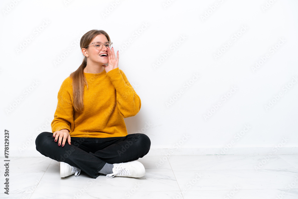 Young caucasian woman sitting on the floor isolated on white background shouting with mouth wide open to the lateral Young caucasian woman sitting on the floor isolated on white background shouting with mouth wide open to the lateral
