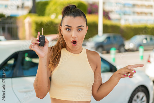Young pretty woman holding car keys at outdoors surprised and pointing side