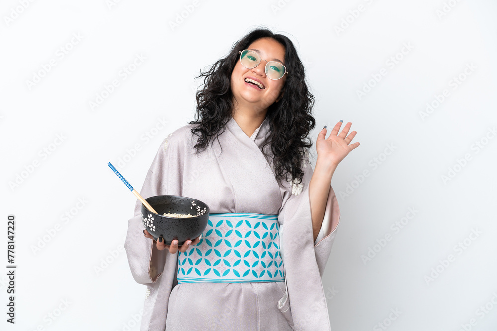 Young asian woman wearing kimono isolated on white background saluting with hand with happy expression while holding a bowl of noodles with chopsticks Young asian woman wearing kimono isolated on white background saluting with hand with happy expression while holding a bowl of noodles with chopsticks