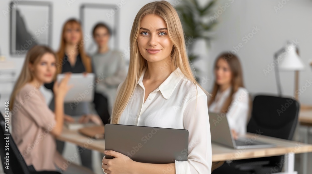 Obraz premium Portrait of attractive female entrepreneur in casual clothes holding laptop in hand standing in office, looking at camera. Startup business team discussing project sitting at desk on background.