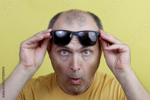 Studio shot of a man 40-50 years old, holding black glasses, looking surprised and funny, wearing a yellow T-shirt, against a yellow studio background.