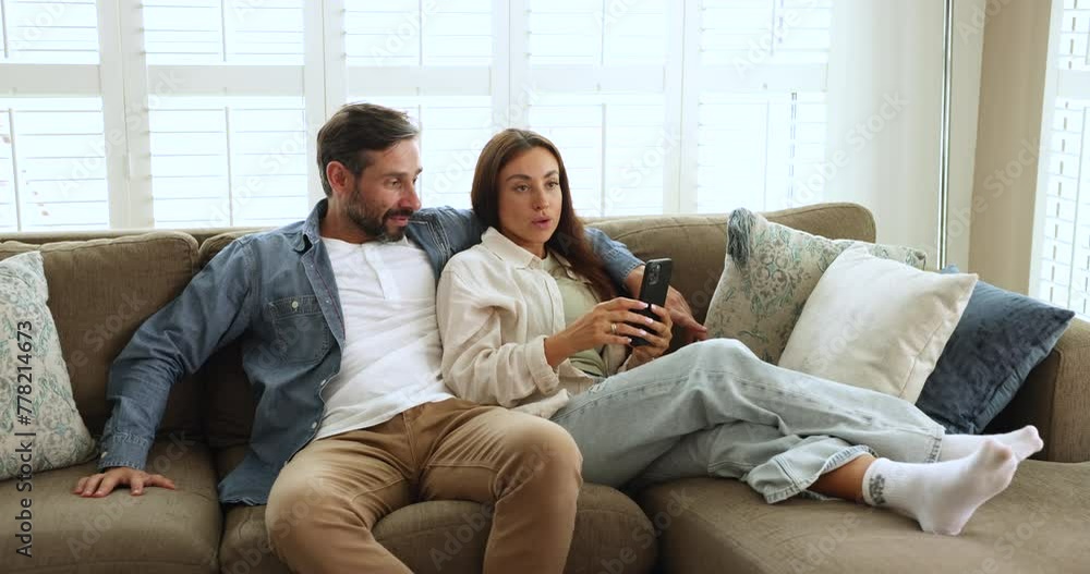 Couple relaxing on couch with smartphone, smile, enjoy talk, discuss fresh news, having conversation about new mobile application, spend carefree leisure together at home. Pastime, modern tech usage Couple relaxing on couch with smartphone, smile, enjoy talk, discuss fresh news, having conversation about new mobile application, spend carefree leisure together at home. Pastime, modern tech usage