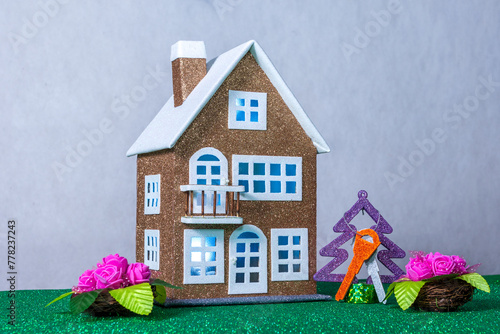 Near a brown toy house with blue light in the windows there are flowers and a Christmas tree with keys