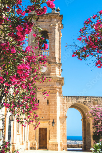 Picturesque photograph of a stone church with pink oleander flowers in the foreground and bright blue skies in the background. Location is Mellieha, Malta. photo