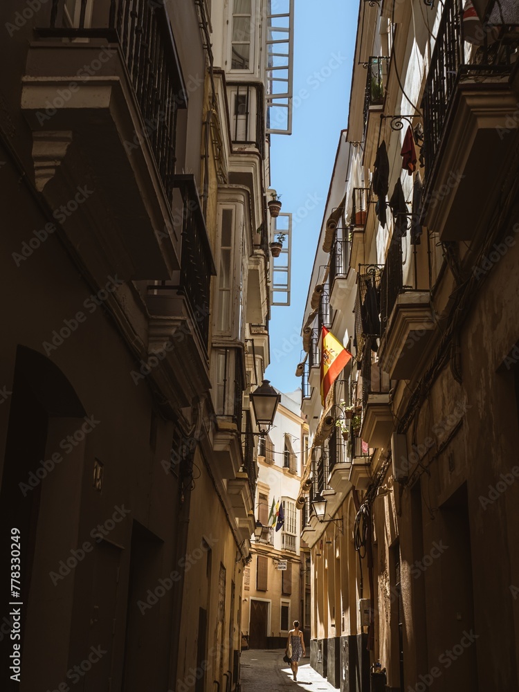 A young woman tourist in a blue dress walking down a narrow old town street in Cadiz under a Spanish flag, Andalusia, Spain