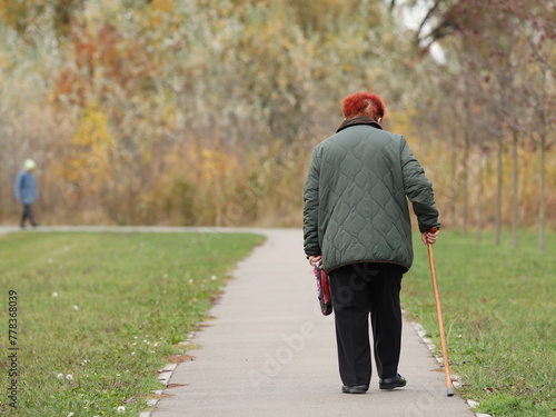 A lonely elderly woman with red hair walks with a stick along the alley of an autumn park. Breathe fresh air in nature. Active aging. Healthy lifestyle in old age