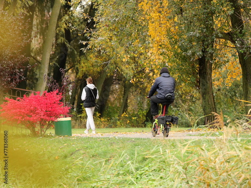 A man on a city electric bicycle catches up with a girl on the alley of an autumn park. Healthy and active lifestyle in the city. Romantic holiday in nature. Traveling on two wheels