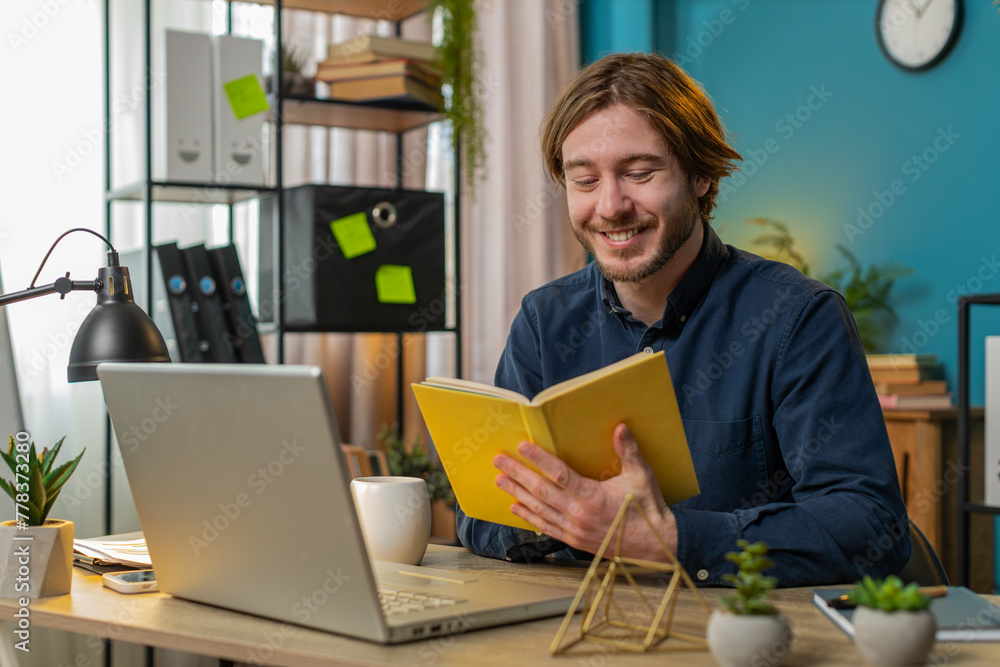 Caucasian young businessman reading interesting book, turning pages smiling enjoying literature, taking a rest after work. Happy cheerful freelancer guy with laptop relaxing at home office table. Caucasian young businessman reading interesting book, turning pages smiling enjoying literature, taking a rest after work. Happy cheerful freelancer guy with laptop relaxing at home office table.