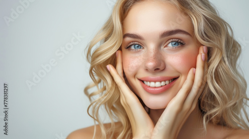 A young European blonde woman with a joyful expression against a white background. Woman with natural makeup  showcasing radiant skin and a bright smile. Curly hair frames her face  hands on cheeks
