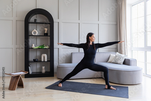 Athletic brunette practicing asanas at home, doing exercise Virabhadrasana 1, warrior pose number one, practicing yoga in black sports overalls standing on a mat, full-length view