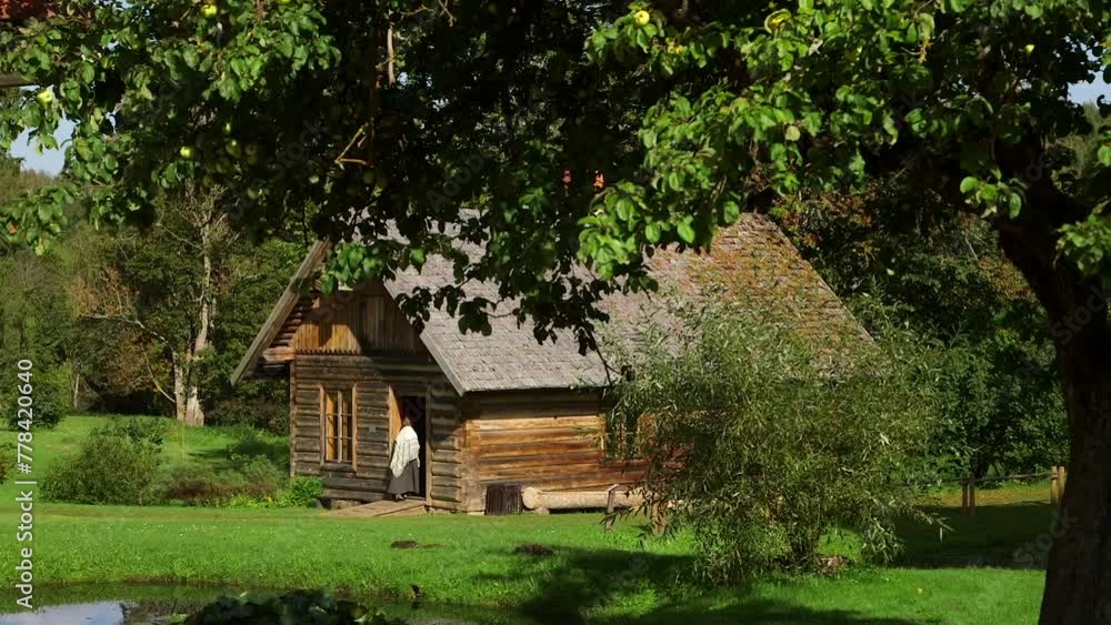 Woman in Historical Costume Enters the Ancient Log House. A Log House, or Log Building, is a Structure Built With Horizontal Logs Interlocked at the Corners by Notching. Woman in Historical Costume Enters the Ancient Log House. A Log House, or Log Building, is a Structure Built With Horizontal Logs Interlocked at the Corners by Notching.