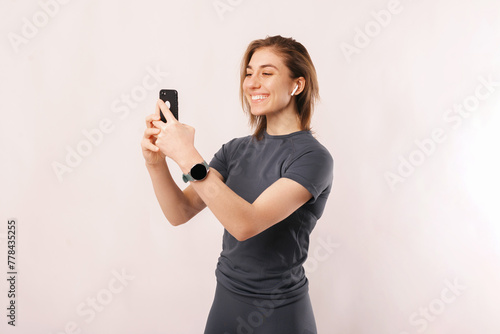 Studio portrait of young smiling woman in sportswear using phone and ear buds. She takes a selfie or having a video call.