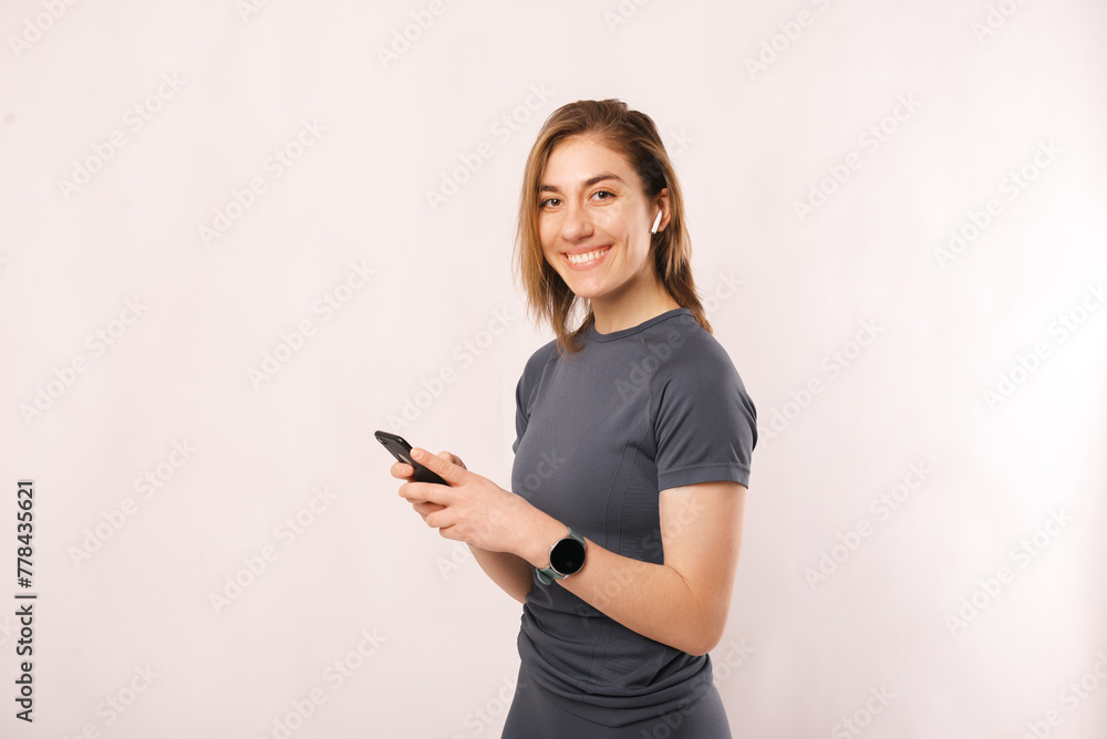 Studio portrait of young smiling woman in sportswear using phone and ear buds over white background. Studio portrait of young smiling woman in sportswear using phone and ear buds over white background.