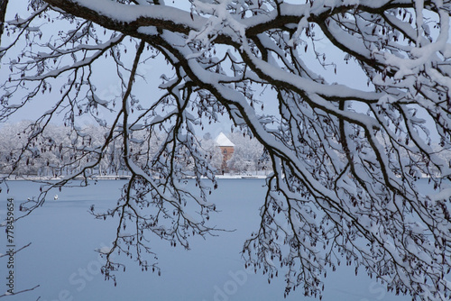 The beauty of a winter's day is on display in this image of Bernardine Lake in Trakai, Lithuania. The sun shines brightly overhead, casting a warm glow on the snow-covered landscape.