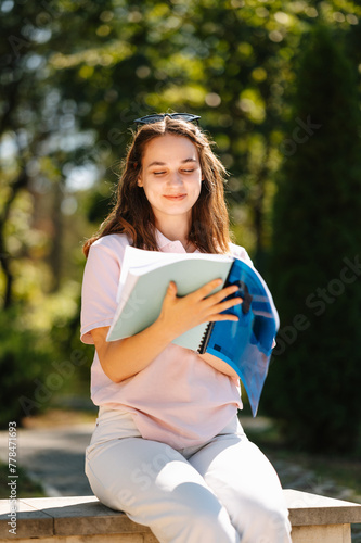 Young smiling female student is sitting outdoors reading on a sunny day in park.