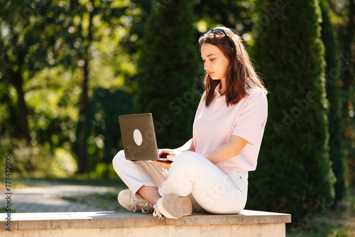 Young concentrated woman is sitting in park using laptop for her studies on a sunny day.