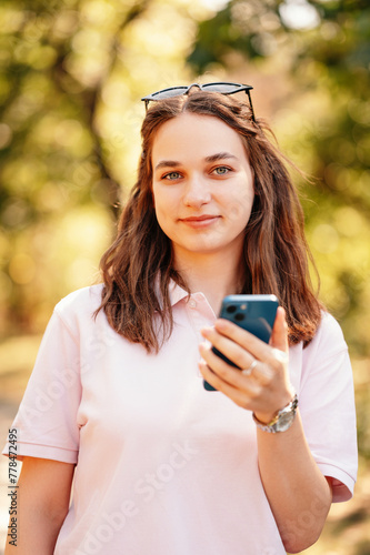 Close up vertical shot of a smiling woman while holding her phone outdoors in park.