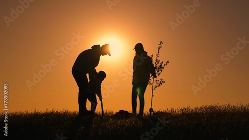 Family with tree at sunset Silhouette. Mother father child team planting tree in sun spring time. Family with shovel  watering can plants young trees sprout in soil. Farmer dad mom child planting tree