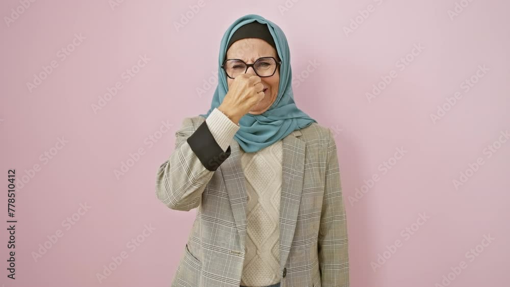 Middle-aged hispanic woman in hijab disgusted by intolerable stink, holding nose over stinky smell, isolated on pink background