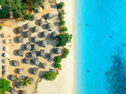 Aerial view of green palm trees, umbrellas on the empty sandy beach, blue sea at sunset. Summer travel in Nungwi, Zanzibar island. Tropical landscape with palms, white sand, clear ocean. Top view