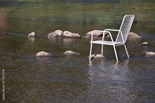 In summer, a white metal chair stands in the middle of a river. There are stones in the middle of the water. The sun is shining. There is space for text.