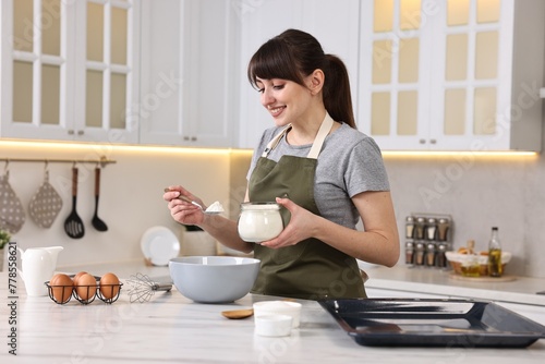 Happy young housewife adding flour into bowl at white marble table in kitchen. Cooking process