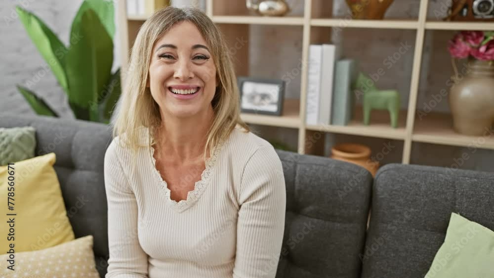 Optimistic middle age blonde woman sitting confidently on her living room sofa, smiling joyfully while pointing to the side with a friendly hand gesture at home. Optimistic middle age blonde woman sitting confidently on her living room sofa, smiling joyfully while pointing to the side with a friendly hand gesture at home.