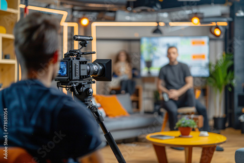 a man being interviewed in front of the camera, sitting on sofa and talking to host while behind him there is an professional video engineer shooting with his cinema camera on tripod 