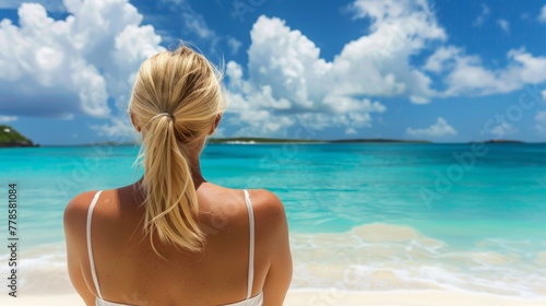 A blonde woman in a white bikini standing on a beach, gazing out at the ocean © tashechka