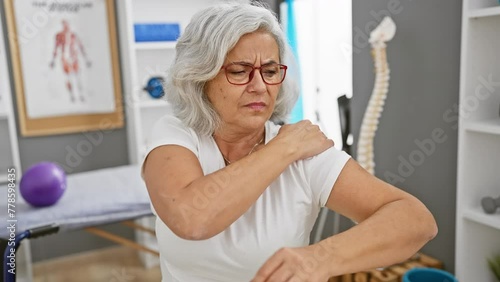 Grey-haired woman in glasses experiencing shoulder pain in a physiotherapy clinic interior. photo